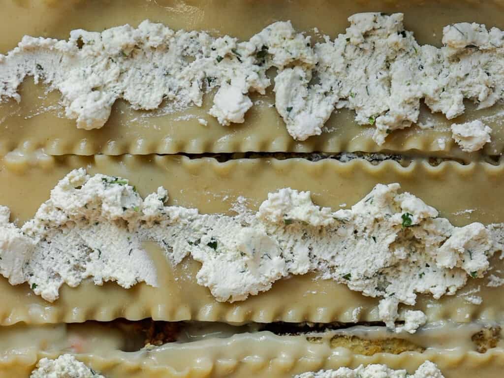 Close-up of uncooked lasagna noodles with dollops of ricotta cheese spread evenly on top. The ricotta is mixed with herbs, adding a speckled green appearance to the white cheese. The texture of the noodles and cheese is clearly visible.